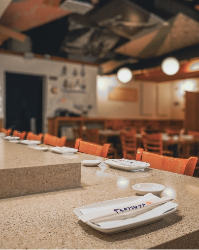 Close-up of a sushi bar with white plates and chopsticks on a granite counter; orange chairs are visible in the background.