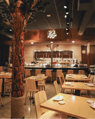 Modern Japanese restaurant dining room with light wood tables and chairs, a decorative indoor tree, and a sign with the 'Katsu' character.