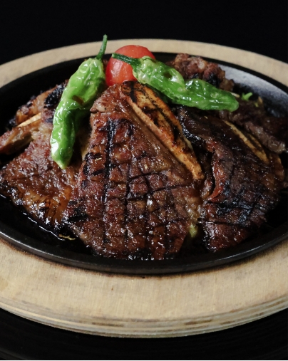 Close-up of sizzling, glazed short ribs on a cast iron platter, garnished with bright green shishito peppers and a cherry tomato.
