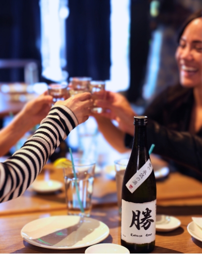 Customers cheering with small sake glasses over a wooden table; a bottle of Katsuya sake is visible in the foreground.