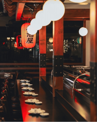 Dark, intimate interior of a Japanese restaurant showing a long counter with small serving plates and decorative white and red lanterns.