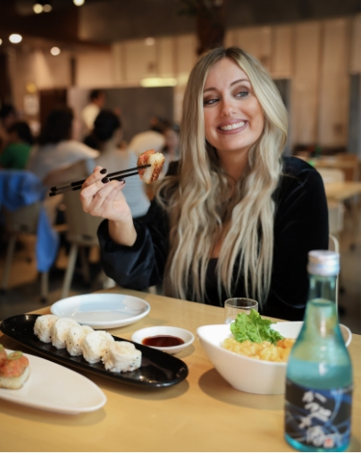 A woman smiling while holding a piece of sushi with chopsticks, with a plate of sushi and a bottle of sake on the table in the foreground.