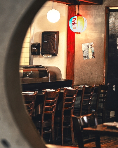 Japanese restaurant sushi counter and high chairs, viewed through a circular opening, with an illuminated Orion beer lantern hanging overhead.