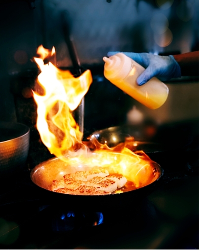 Chef in blue gloves pouring oil into a pan to create a large flame while searing rice patties in a commercial kitchen.
